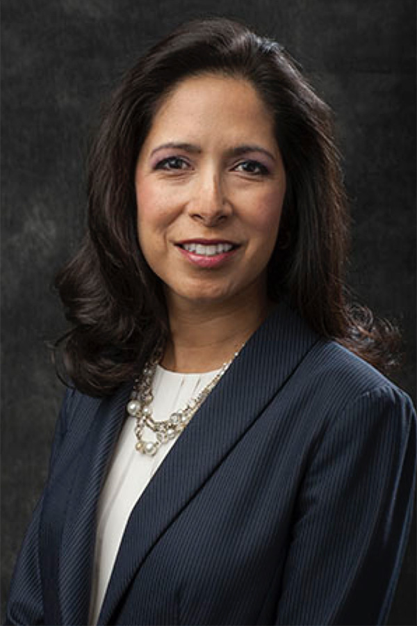 Woman with long dark hair in a navy blazer and white blouse, wearing a statement necklace, smiles in front of a dark background.