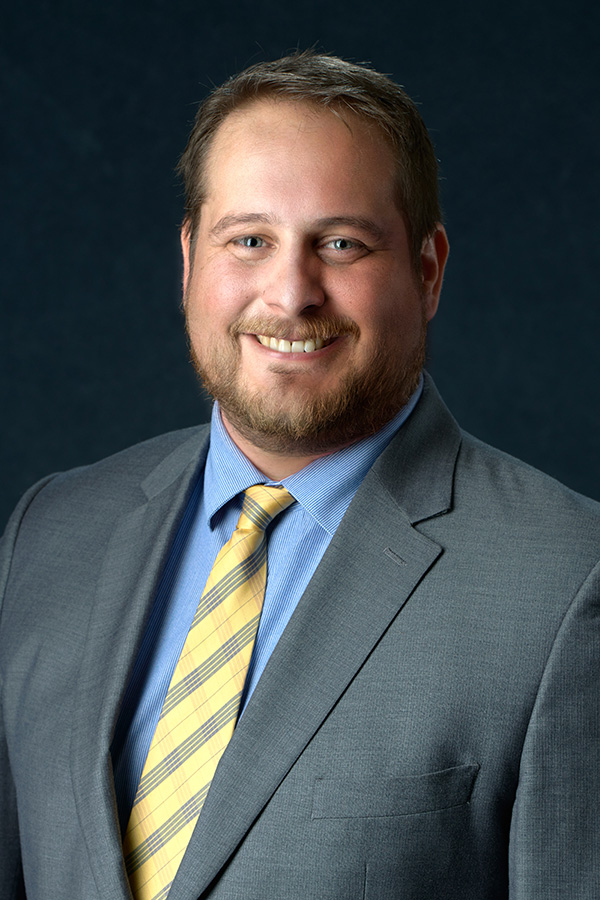 A man with short hair and a beard wearing a gray suit, blue shirt, and yellow striped tie, smiling against a dark background.