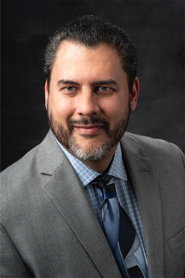 Man with dark hair and beard wearing a grey suit, blue patterned shirt, and blue tie poses for a professional portrait against a dark background.