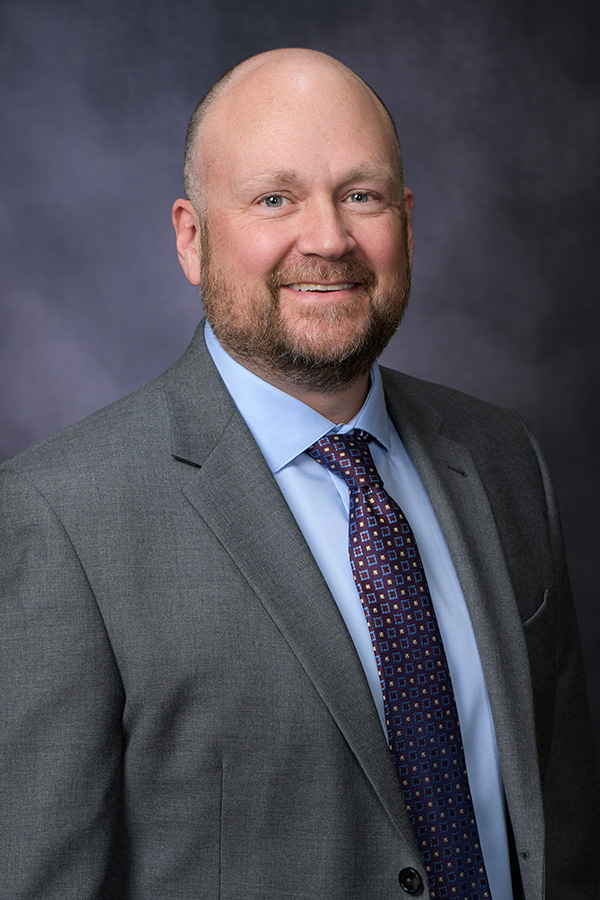 A man in a gray suit, blue shirt, and patterned tie poses in front of a dark studio background, smiling at the camera.