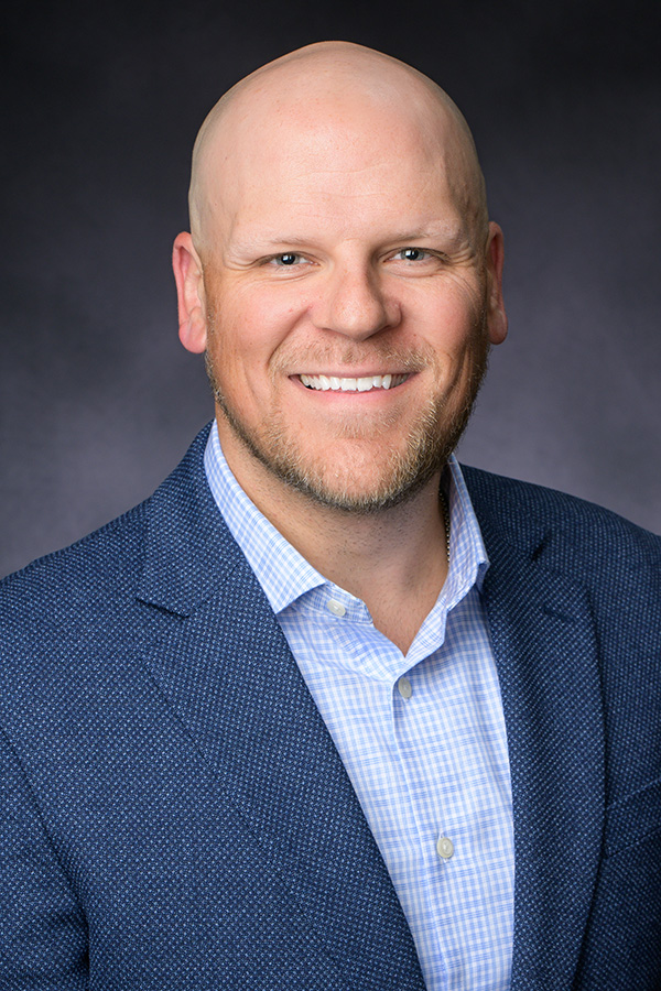 A smiling man with a bald head, wearing a blue textured blazer over a light blue checkered shirt, poses against a dark, plain background.