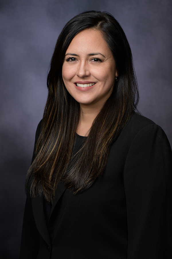 Woman with long dark hair wearing a black blazer, standing in front of a dark, blurred background, smiling at the camera.