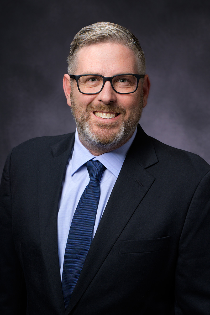 A man with short gray hair, a beard, and glasses is wearing a dark suit, light blue shirt, and navy tie, smiling in front of a dark, neutral background.
