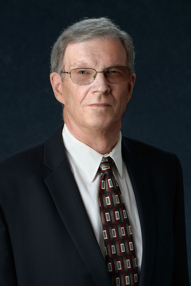 An older man with gray hair and glasses wearing a dark suit, white shirt, and patterned tie, posing against a dark background.