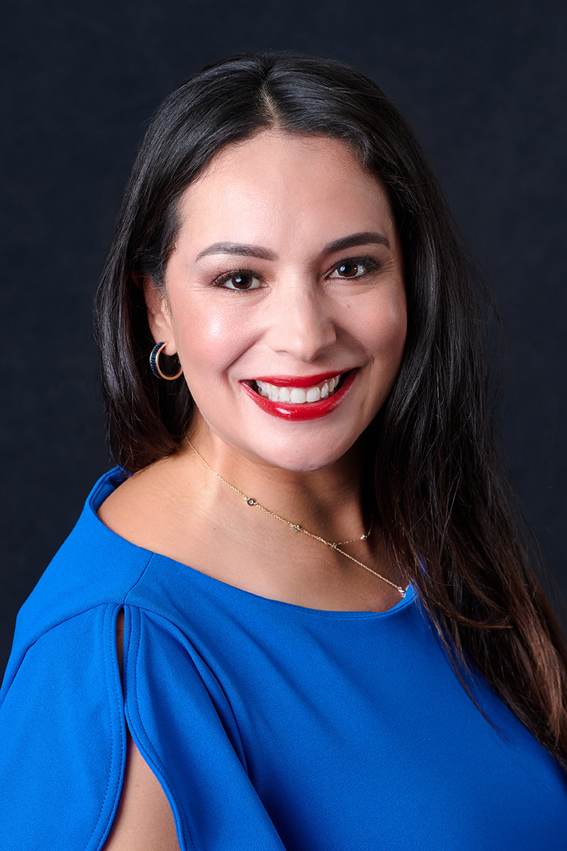 Woman with long dark hair, wearing a bright blue top and gold hoop earrings, smiling in front of a plain dark background.