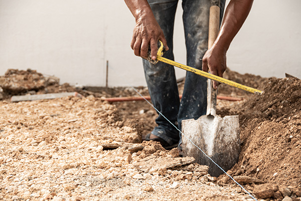 Person measuring width of a trench with a tape measure while holding a shovel, surrounded by dirt and gravel at a construction site.