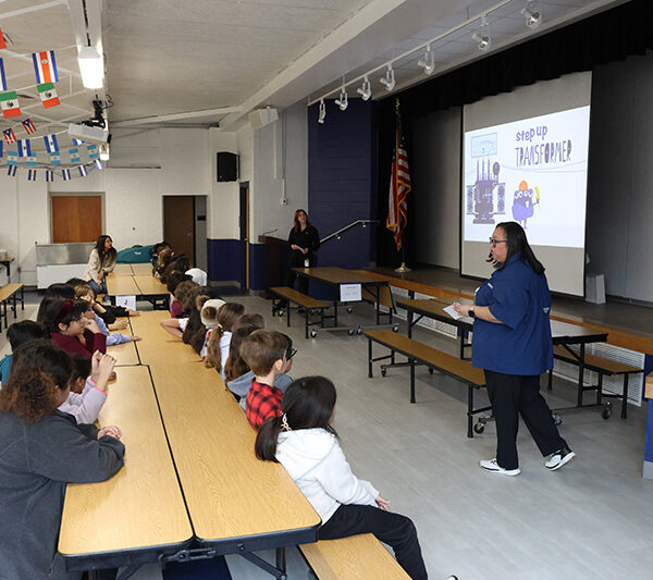 A woman stands in front of a group of students seated at cafeteria tables, presenting a slideshow titled 