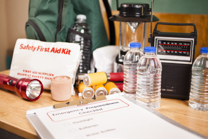 Emergency preparedness checklist on clipboard and a large pile of supplies to be used in case of a natural disaster (hurricane, flood, earthquake, etc.) including: flashlight, batteries, water bottles, first aid kit, lantern, radio, backpack, mask. No people.